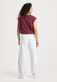 Femme avec des cheveux bouclés portant un haut sans manches bordeaux et un pantalon blanc ample, se tenant debout et tournant le dos contre un fond uni.