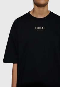 Svart bomullst-shirt med korta ärmar, med en central tryckt logotyp "HALO" i beige, tillsammans med numeriska detaljer under logotypen.