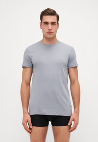 T-shirt gris à manches courtes en tissu doux, avec un col rond et une silhouette ajustée. Porté avec un short noir.