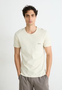 T-shirt en coton beige clair avec un col rond et des manches courtes. Présente un petit logo vert "BOSS" sur la poitrine gauche.