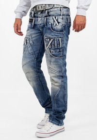 Jean en denim bleu clair avec plusieurs poches, coutures renforcées et finition délavée. Associé à des baskets blanches et un sweat-shirt gris.