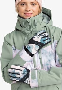 Une veste imperméable verte avec un motif de paysage pastel, complétée par des gants noirs et à motifs arborant le logo Roxy.
