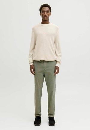 Pull en tissu crème à texture avec des manches longues, associé à un pantalon slim vert clair et des chaussures à lacets noires. Tenue décontractée simple et moderne.