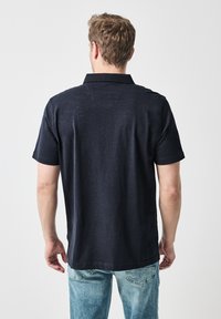 Marinblå kortärmad polo i lätt tyg med krage och rak fåll, som visar subtil textur och sömnadsdetaljer.