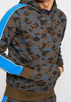 Hombre con una sudadera con capucha de patrón de camuflaje con rayas azules y blancas en las mangas y las manos en el bolsillo delantero.