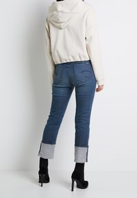 Skuren krämfärgad hoodie, figursydda blå jeans med grå manschetter, dragkedjedetaljer på fickorna och svarta högklackade stövlar. Släta tygtexturer.