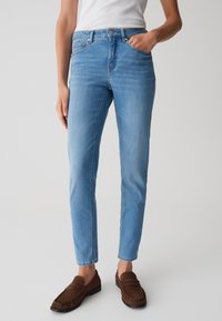 Jean en denim bleu clair avec une coupe ajustée, design à cinq poches et fermeture à bouton en métal. Assorti à des mocassins en cuir suédé marron.