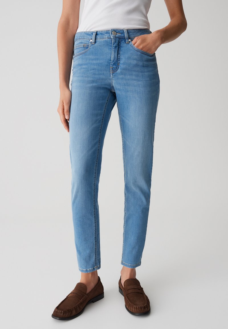 Jean en denim bleu clair avec une coupe ajustée, design à cinq poches et fermeture à bouton en métal. Assorti à des mocassins en cuir suédé marron.