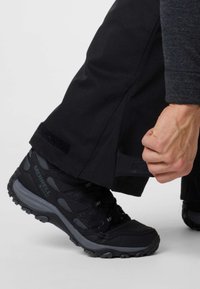 Black hiking boot with textured details and gray accents, paired with black pants featuring a Velcro adjustment at the hem.