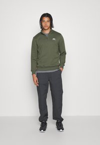 Felpa verde scuro con zip, t-shirt grigia sotto, abbinata a pantaloni larghi grigi e sneakers nere. Design semplice con dettaglio del logo.