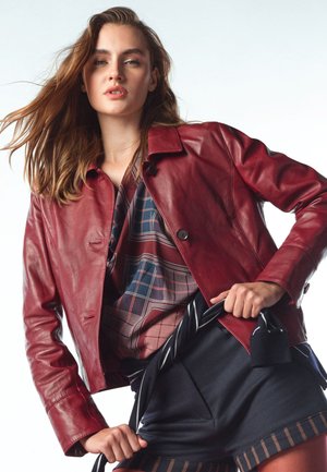 Młoda kobieta z długimi włosami, ubrana w czerwoną skórzaną kurtkę na wzorzystej koszuli, przeciągająca paski na pasku w paski związanym w talii.