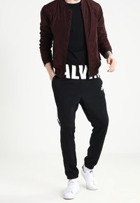 Person klädd i en bordeauxfärgad bomberjacka, svart Calvin Klein-tröja, svarta Nike-joggers med vit rand, och vita Nike-sneakers, står avslappnat.