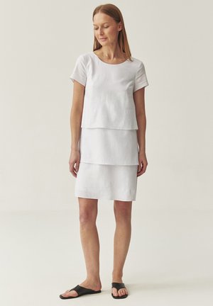 Vestido de lino blanco con diseño en capas; mangas cortas, escote redondo y a la altura de la rodilla. Llevado con sandalias de slide negras. Textura suave.