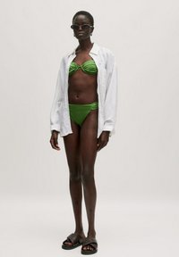 Groene bikini met een gestructureerde afwerking, witte oversized blouse, en donkere slip-on sandalen. Model staat tegen een effen achtergrond.