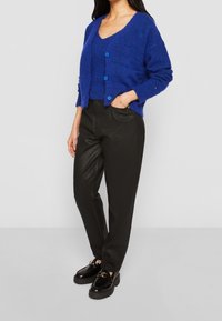 Blå, strukturerad kardiagan med blå knappar lagd över en matchande tröja. Svarta, högmidjade, konstläderbyxor. Svarta platåsandaletter.