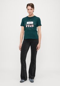 T-shirt verde escuro com uma gráfica de "Feliz Natal" e design de pinguim, combinado com calças flare pretas brilhantes e ténis cinzas.