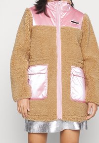 Faux fur-jacka i tan med glänsande rosa detaljer. Har en dragkedja fram, hög krage och två framfickor med rosa foder. Silverkjol under.