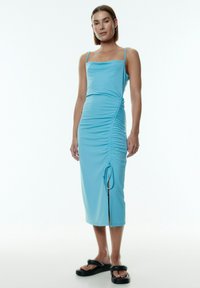 Himmelblaues Midikleid mit quadratischem Ausschnitt, verstellbarem seitlichen Raffung und einem Oberschenkel-spalt. Glattes, dehnbares Material; getragen mit schwarzen Sandalen.
