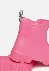 Ružové gumové členkové čižmy s textúrovanými podrážkami a farebným ťaháčikom. Majú hladký vonkajší povrch a pevný, zaoblený dizajn.