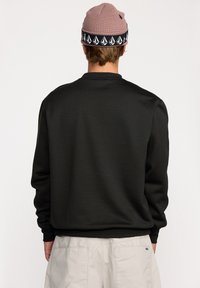 Sweat-shirt noir à manches longues, avec des poignets côtelés et une texture lisse. Porté avec un bonnet en tricot rose orné d'une bande noir et blanc.