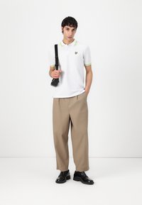 Polo bianco con dettagli verdi, pantaloni beige oversize e scarpe nere. La maglietta presenta un piccolo logo nero sul petto.