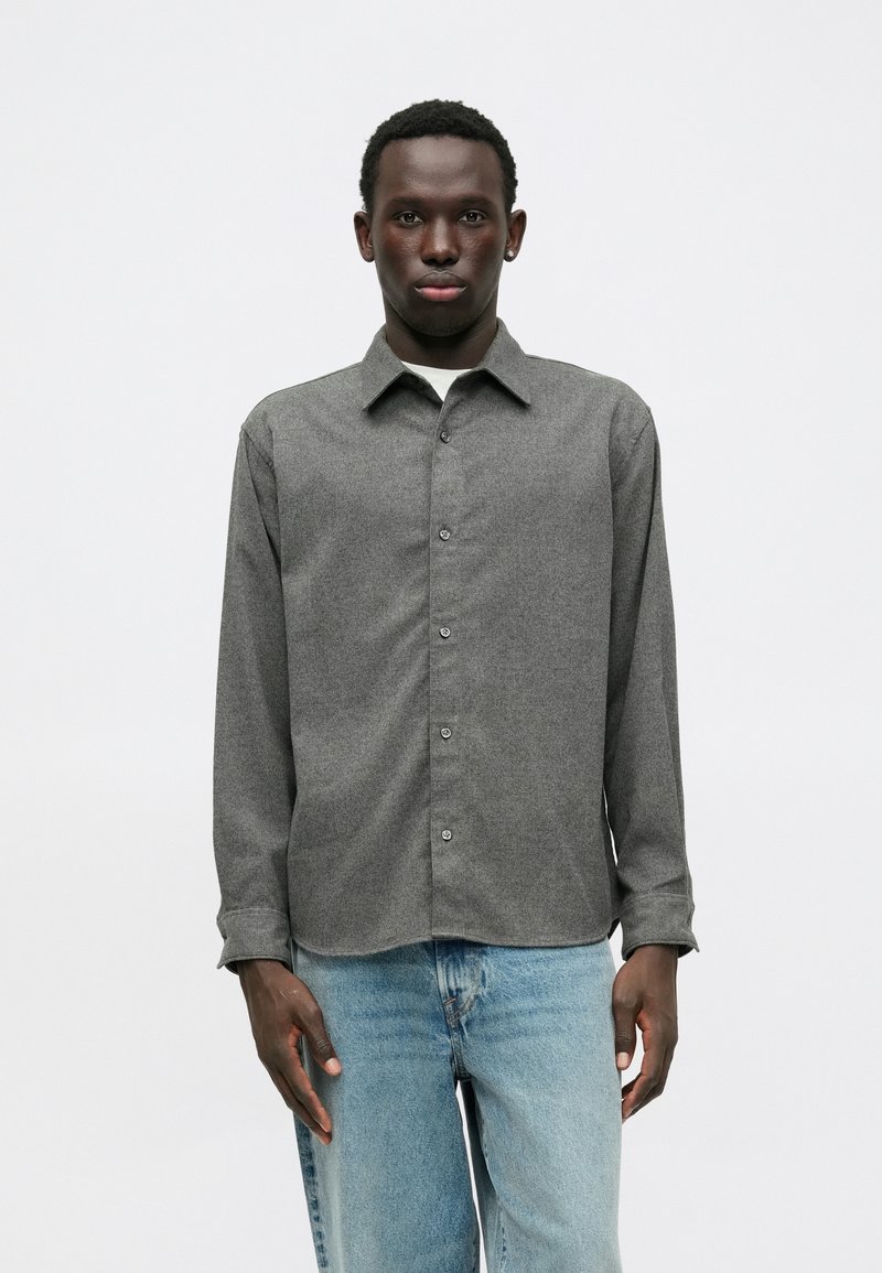 Chemise grise à boutons, fabriquée en tissu texturé, avec un col classique, des manches longues et une patte de boutonnage à l'avant avec des boutons contrastants.