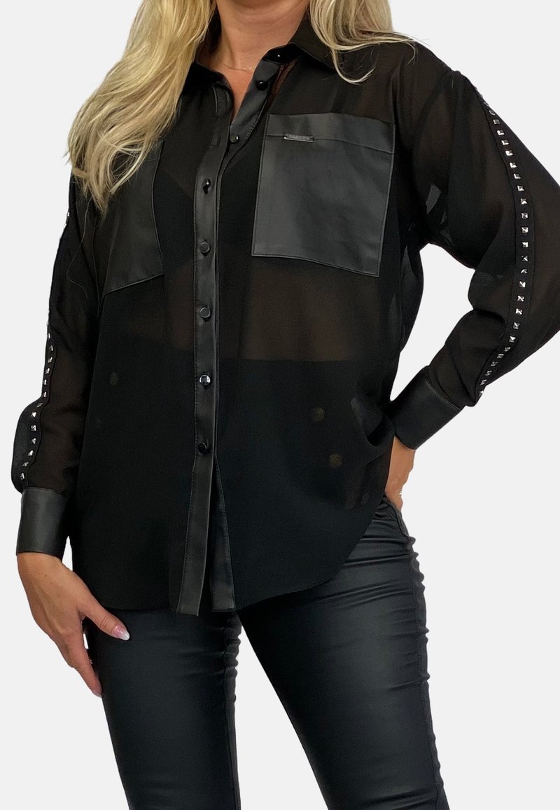 Femme portant une blouse noire transparente à boutons avec des détails en cuir et des manches cloutées, associée à un pantalon en cuir noir, posant devant un fond uni.