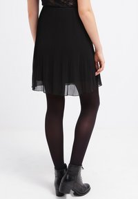 Jupe plissée noire en tissu léger, avec une coupe fluide. Portée avec des collants opaques noirs et des bottines, vue de dos.