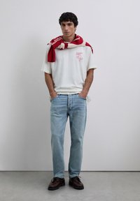 Młody mężczyzna ubrany w białą koszulkę z grafiką, jasno niebieskie dżinsy, ciemno brązowe buty oraz sweter w czerwono-białe paski zarzucony na ramiona, stojący.