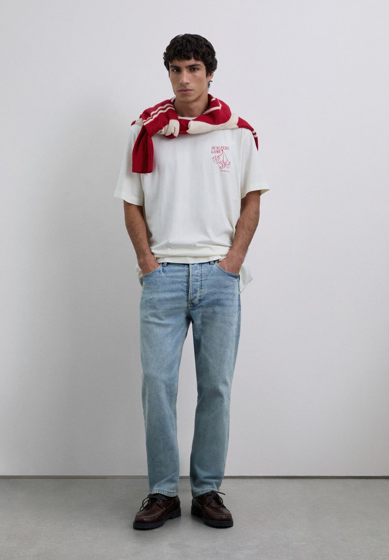 Junger Mann in einem weißen T-Shirt mit Grafik, hellblauen Jeans, dunkelbraunen Schuhen und einem rot-weißen gestreiften Pullover, der über den Schultern drapiert ist, steht.