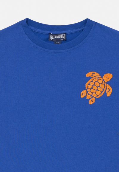 Sinine ümara kaelusega T-särk, millel on üleval vasakul rinnal tikitud oranž kilpkonnamotiiv ning Vilebrequin kaubamärgi silt krae sees.