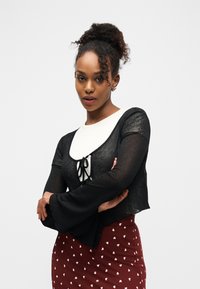 Νεαρή γυναίκα με κατσαρό ψηλό χτένισμα φορώντας μαύρο διαφανές τοπ πάνω από λευκό πουκάμισο και κόκκινη φούστα με λευκά σχέδια καρδιών, με σταυρωμένα τα χέρια.