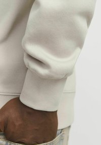 Lichtbeige sweatshirt met geribbelde manchetten. Heeft een zachte stof en een relaxte pasvorm. Een hand is in de zak van spijkerbroek geplaatst.