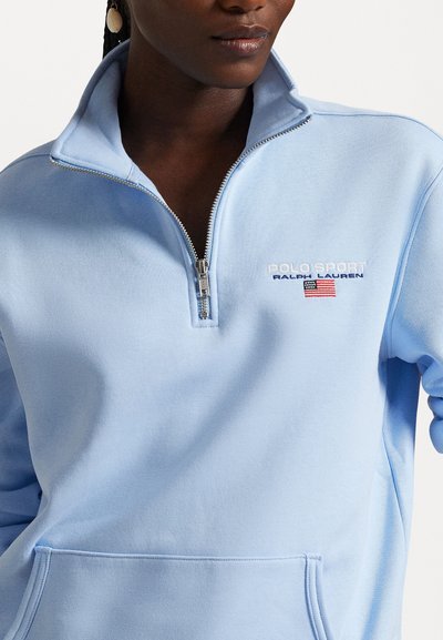 Jasnoniebieski sweter, zapinany na zamek, z wysokim kołnierzem, z haftem "POLO SPORT" oraz amerykańską flagą w białym i czerwonym kolorze. Miękka tkanina.