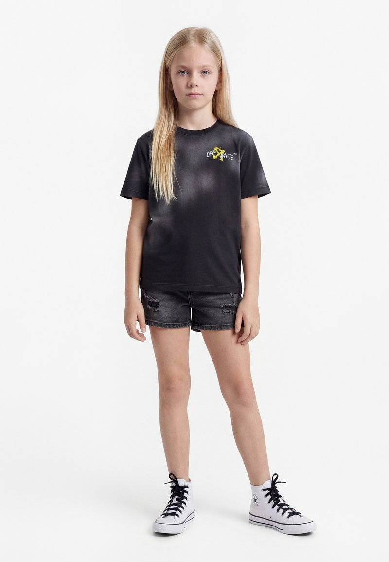 Blondes Mädchen trägt ein schwarzes Off-White T-Shirt, zerrissene schwarze Jeansshorts und weiße High-Top-Sneaker mit schwarzen Schnürsenkeln und steht vor weißem Hintergrund.