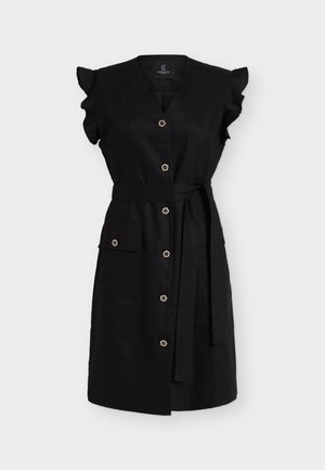 Femme aux cheveux blonds courts portant une robe noire boutonnée avec des manches à volants et une ceinture, tenant une pochette dorée, et des talons hauts noirs.