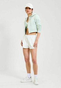 Sudadera corta en verde menta, sujetador deportivo en verde claro, pantalones cortos blancos, zapatillas blancas y una gorra blanca; un look atlético casual con materiales suaves.
