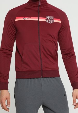 Bordowa kurtka sportowa na zamek błyskawiczny z różowym paskiem, logo Nike i herbem FC Barcelona, noszona z szarymi sportowymi spodniami Nike.
