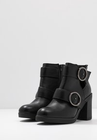 Schwarze Knöchelstiefel aus synthetischem Leder, mit runden Metallakzenten, einem Blockabsatz und seitlichen Reißverschlüssen für ein einfaches An- und Ausziehen.