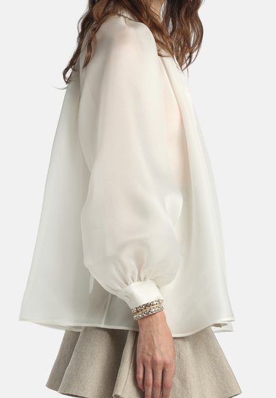 Sieviete valkā caurspīdīgu baltu blūzi ar garām piedurknēm ar sapinētiem manžetēm un bēšā krāsā plīša svārku, redzama sānskats no pleciem līdz gurniem.