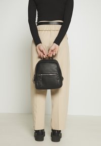 Sac à dos en cuir noir avec une surface texturée, une poignée supérieure et une poche zippée à l'avant. Le modèle porte un pantalon beige et des bottines noires.