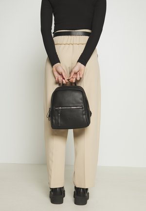 Black leather backpack with textured surface, top handle, and front zip pocket. The model wears beige trousers and black ankle boots.