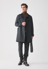 Abrigo trench coat azul oscuro con diseño de doble botonadura, botones negros, llevado sobre un suéter de cuello alto marrón y pantalones negros, combinado con botas negras.