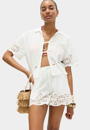 Donna che indossa un completo corto in pizzo sangallo bianco con camicia annodata sul davanti e pantaloncini, tiene in mano una borsa a secchiello color cuoio con frange, indossa braccialetti e orecchini a cerchio dorati.