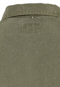 Olijfgroene blouse met een kraag, voorzien van een rechthoekige patch en zichtbare stiksel. De stof lijkt zacht en gestructureerd te zijn.