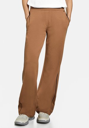 Person trägt lockere braune Hose mit Seitentaschen und schwarzem Paspel, kombiniert mit weißen und grauen Sneakers vor einem einfarbigen Hintergrund.