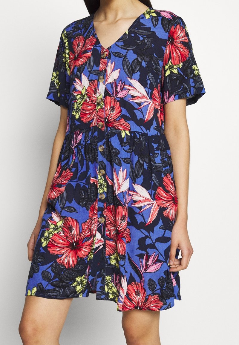 Femme portant une robe bleue à manches courtes avec des motifs floraux rouges et roses et des boutons sur le devant, debout avec une main près de son côté.