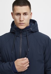 Tamsiai mėlyna striukė su kapišonu ir užtrauktuku, tekstūruota medžiaga ir raištelių akcentais. Modelis laiko užtrauktuką su neutralia fono.