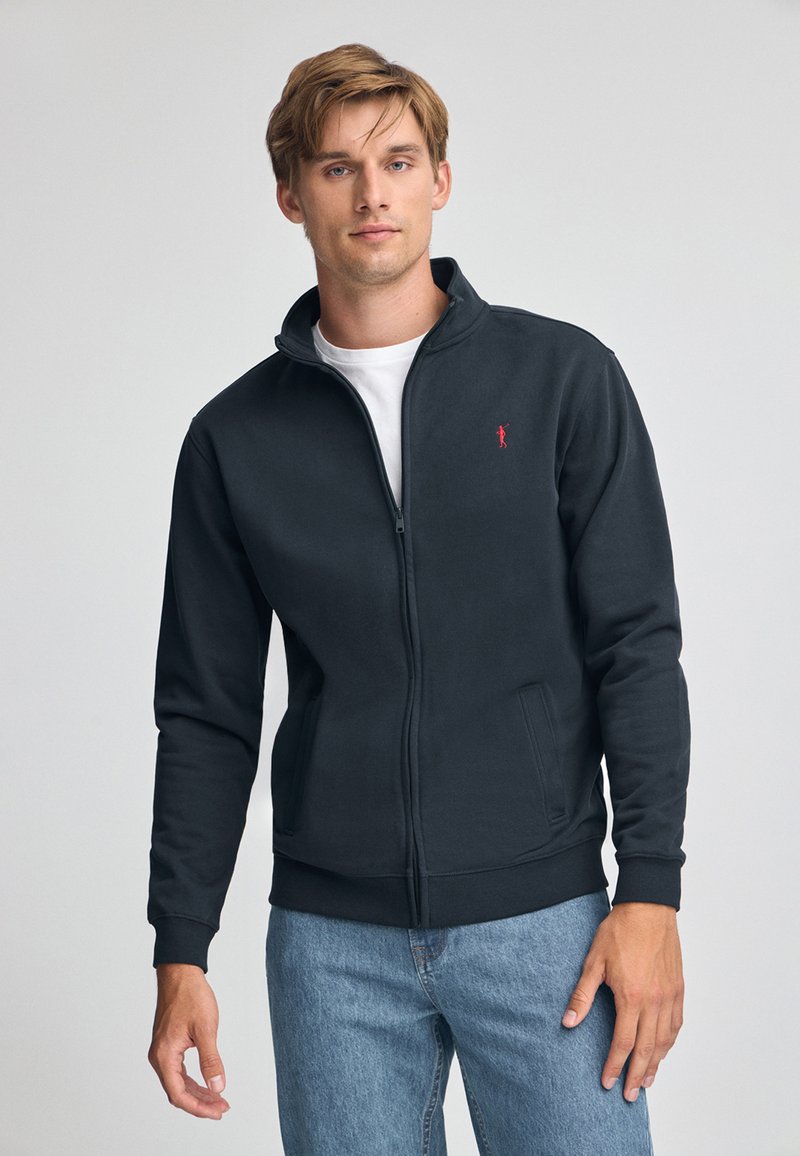 Felpa nera con zip realizzata in materiale morbido, con colletto alto e accento del logo rosso. Abbinata a jeans blu chiaro.