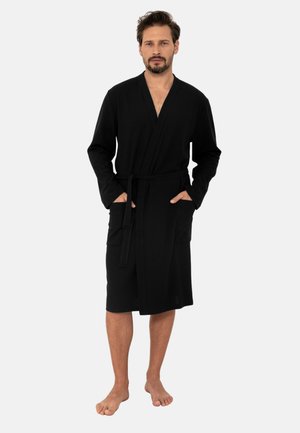 Mężczyzna z brodą i wąsami, ubrany w czarny, sięgający do kolan płaszcz, stoi boso z rękami w kieszeniach na jednolitym tle.